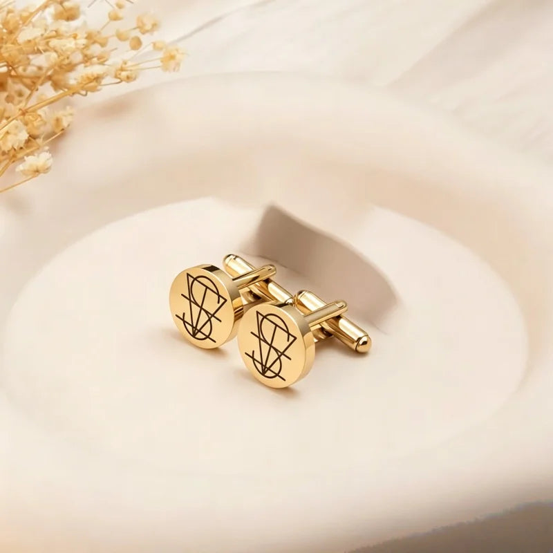 A pair of gold round cufflinks with engraved black monogram designs, displayed on a soft cream-colored dish with dried flowers in the corner.