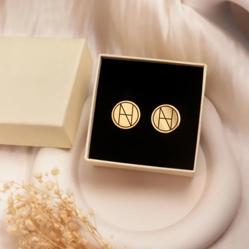 A pair of gold circular stud cufflinks with an interlocking name design, presented in a black square box next to an open cream-colored box on a soft white draped fabric background.