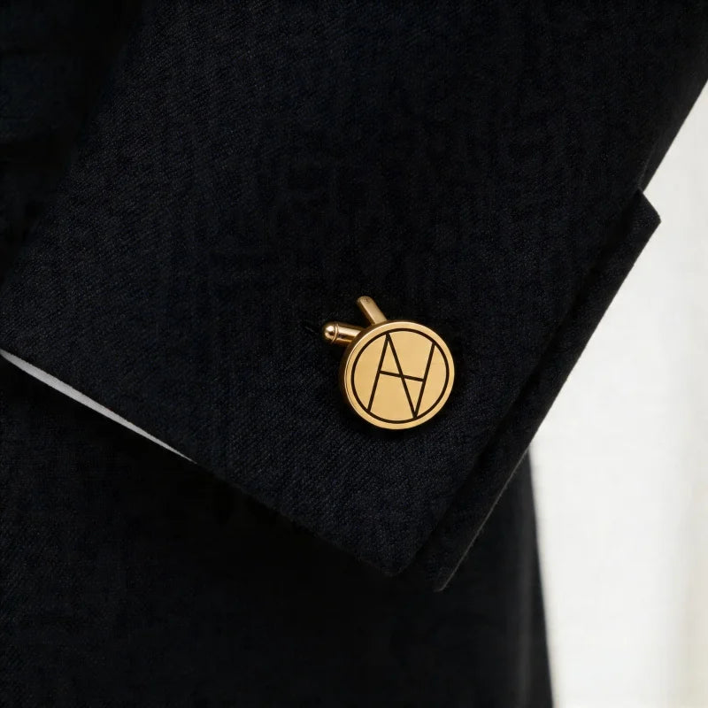 A gold circular cufflink with an interlocking 'A' monogram, pinned to the cuff of a textured black suit jacket.
