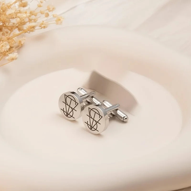 A pair of silver-toned round cufflinks with engraved black abstract line designs, displayed on a light beige ceramic dish with dried flowers in the corner.