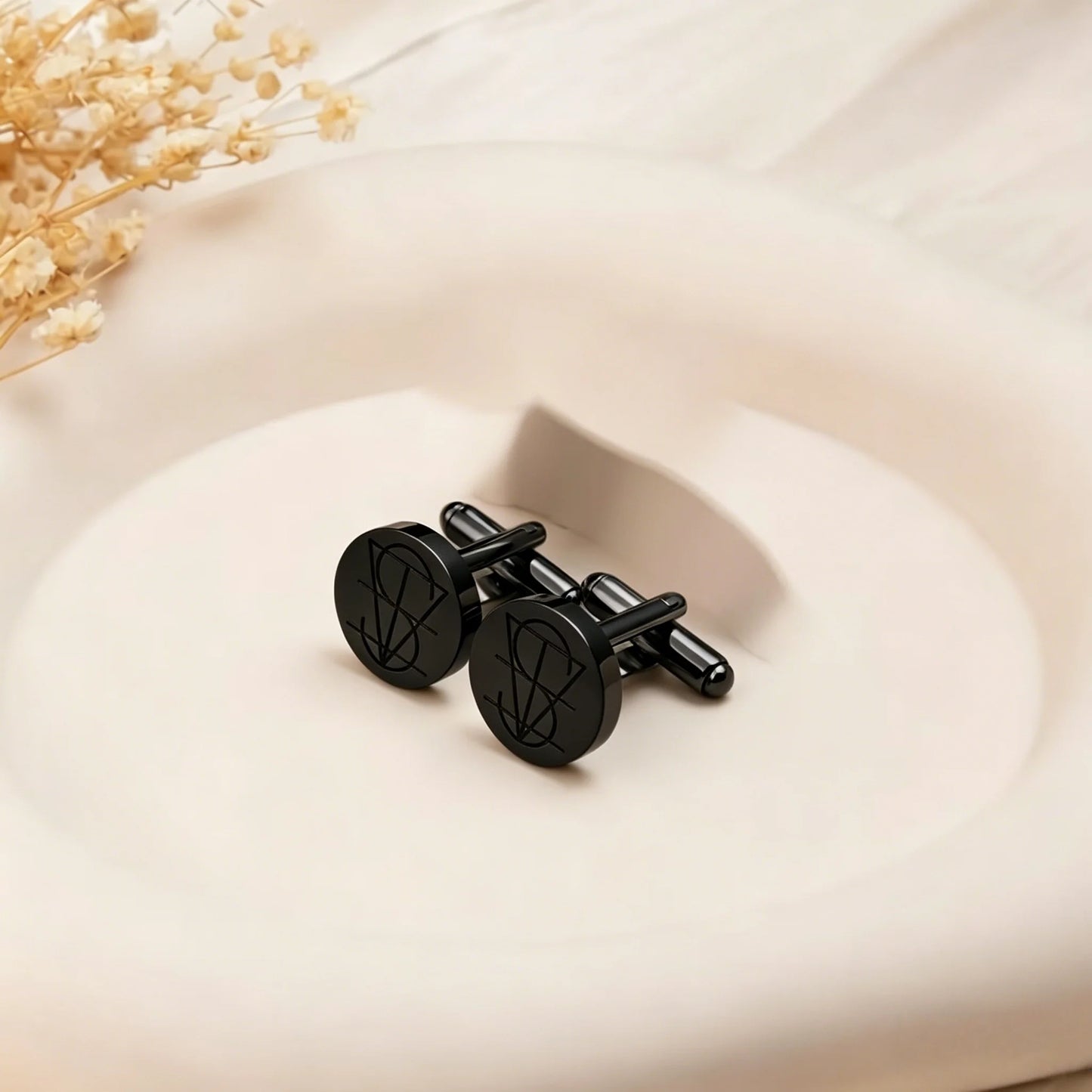 A pair of matte black cufflinks with a pentagram design on the round faces, displayed on a warm beige ceramic dish with dried flowers in the corner.