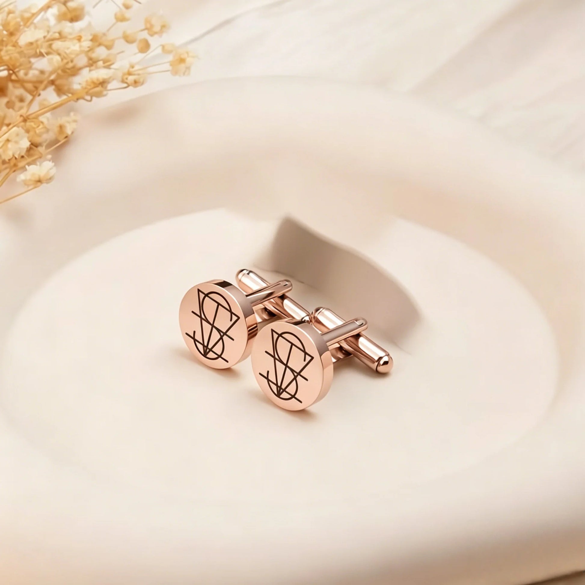 A pair of rose gold cufflinks with engraved black monogram designs, resting on a white ceramic dish with dried flowers in the background.